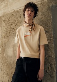 Beige katoenen t-shirt met geborduurd "PUMA" logo; gecombineerd met zwarte cargobroek. Accessoires omvatten een gestreepte beige sjaal. Achtergrond met een gestructureerde muur.