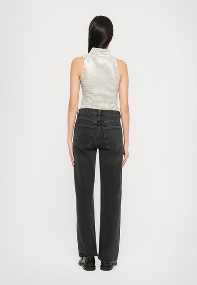 Vrouw met lang, donker haar in een mouwloze gestreepte coltrui, zwarte hoge taille rechte jeans en zwarte schoenen, staande met haar rug naar ons toe.