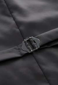 Kangasta valmistatud must kangas, mille pinnal on tekstuur; keskel on ainulaadse kujuga ja detailiga hõbedane metalllukk, mis asub materjali keskel.