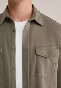 Cămașă verde măslinie cu un aspect neted, având două buzunare pe piept, butoane cu sistem de prindere și guler ascuțit peste un tricou alb.
