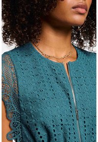 Türkises Spitzenkleid mit aufwendigem Ausschnittmuster, kurzen Ärmeln und einem Frontreißverschluss. Goldene, geschichtete Halsketten sind um den Ausschnitt sichtbar.