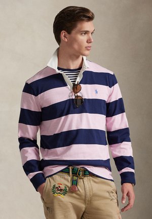 Camisa tipo polo de manga larga a rayas azul marino y rosa con cuello blanco, que presenta un pequeño logo. Se lleva con pantalones beige, adornados con un cinturón colorido.