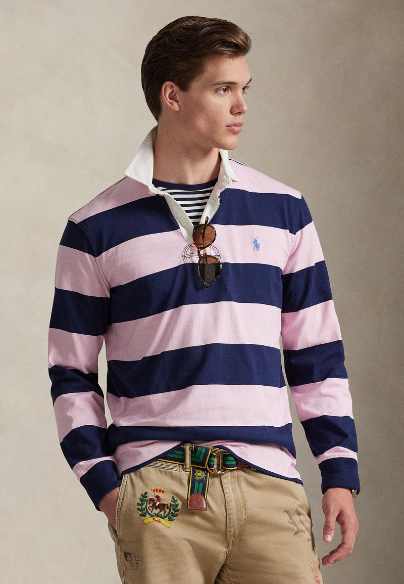 Tumesinise ja roosa triibulise pika varrukaga polo-särk valge kraega, millel on väike logo. Kanditud beežide pükstega, mida kaunistab värvikas vöö.