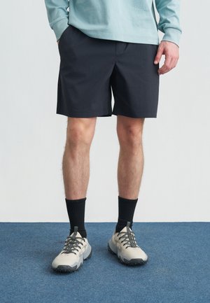 Mand iført sorte shorts, sorte sokker og grå sneakers, står på blåt tæppe med hænderne i lommerne.