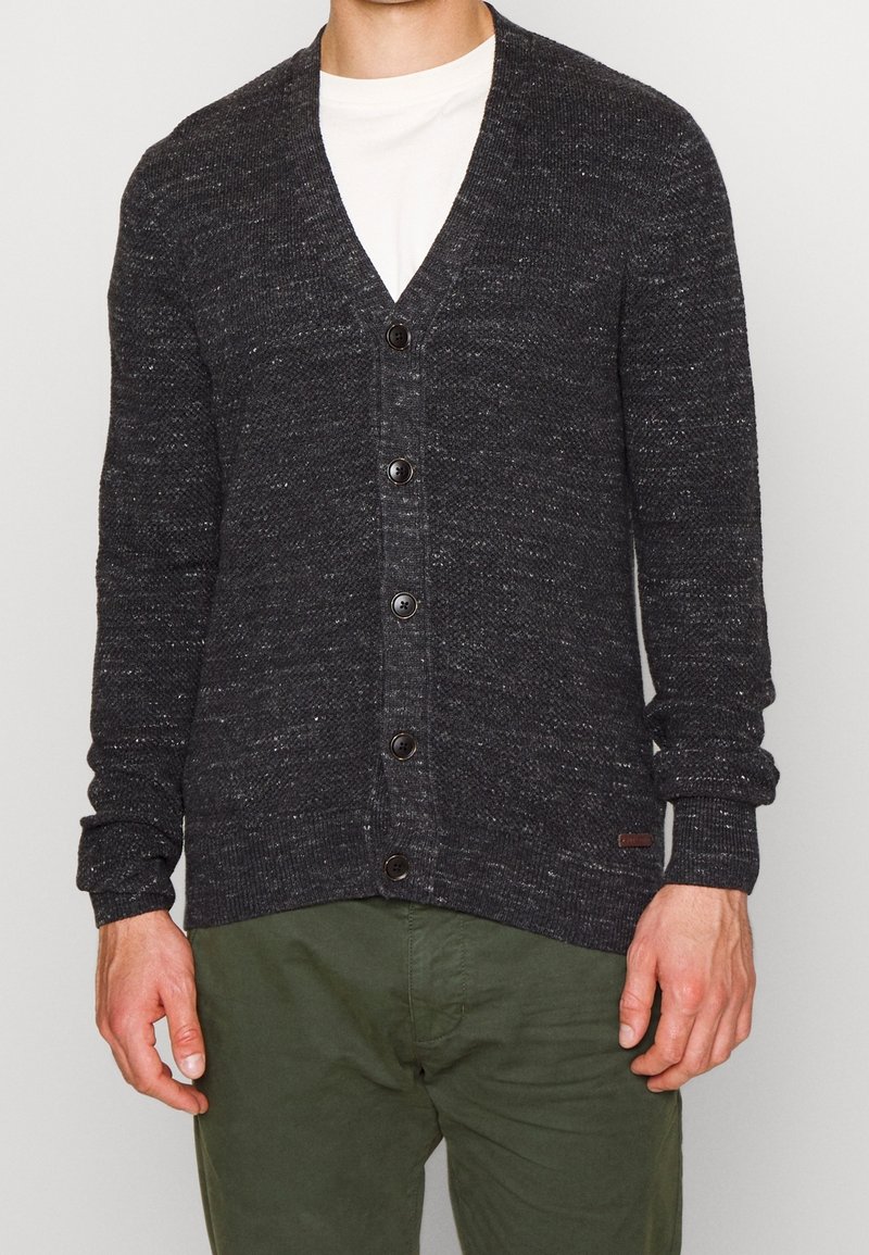 Tmavě šedý pletený kardigan s výstřihem do V, pěti knoflíky, jemně žebrovanými manžetami a strukturovaným vzorem. Nositý přes světlou košili a zelené kalhoty.