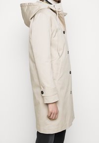 Beige waterdichte jas met capuchon, voorzien van een rechte snit, twee zijzakken, knoopsluitingen en een trekkoorddetail bij de hals.