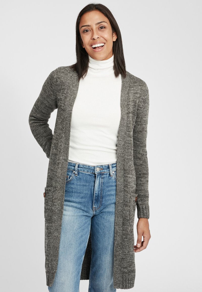 Gray knitted cardigan with a long length, open front, and two front pockets. Worn over a white turtleneck and paired with blue jeans.