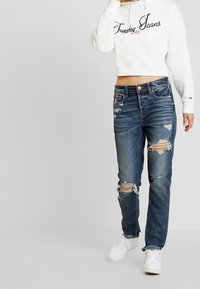 Jeans bleus déchirés avec plusieurs trous et une coupe droite, associés à un hoodie court blanc arborant un logo "Tommy Jeans" en bleu marine.