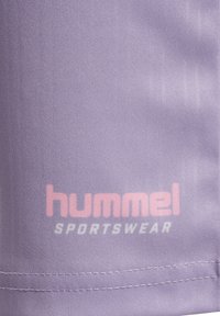 Close-up af lilla sportstøjstof med pink „hummel“ og hvid „SPORTSWEAR“ tekst trykt over syet kant.
