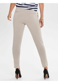 Beige fitted pants with two back pockets, sleek finish, and a high waist, paired with black stiletto heels, showcasing a tailored look.
