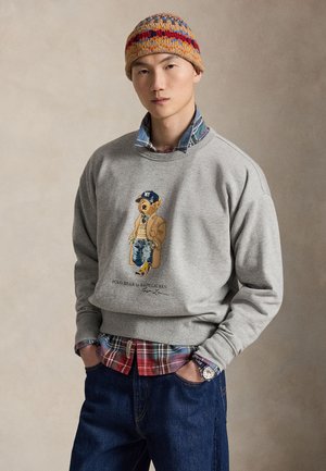Homme portant un bonnet tricoté multicolore, un sweat-shirt gris avec un motif d'ours, une chemise à carreaux en dessous, un jean bleu et une montre-bracelet, posant les mains dans les poches.