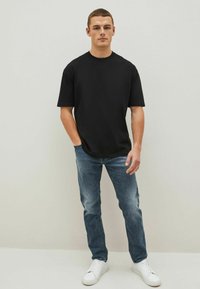T-shirt nera a maniche corte, colletto tondo, abbinata a jeans azzurri e sneakers bianche; il modello si trova davanti a uno sfondo neutro.