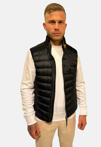Gilet noir en duvet avec fermeture éclair, présentant une texture lisse et un design matelassé, porté sur une chemise blanche à manches longues.