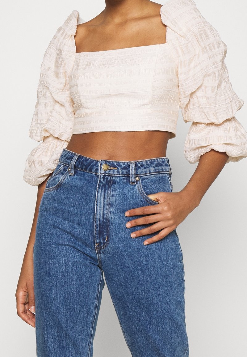 Personne portant un haut court blanc texturé avec des manches bouffantes et un jean bleu taille haute, une main posée sur la hanche.