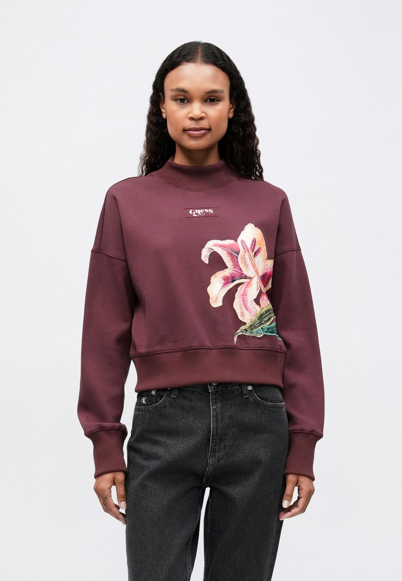 Mujer con cabello rizado largo, viste un sudadera color burdeos con un gran gráfico de un lirio rosa y jeans negros, de pie contra un fondo simple.