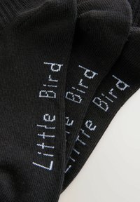 Un par de calcetines negros hechos de un tejido texturizado, con las palabras "Little Bird" bordadas en blanco a lo largo de los lados. Tres pares apilados.