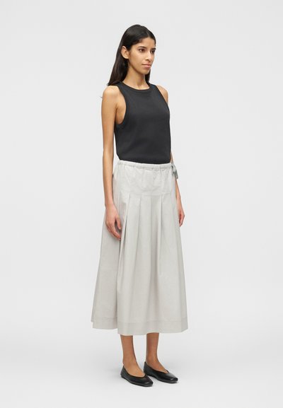 Γυναίκα που φοράει μαύρο αμάνικο τοπ, γκρι ανοιχτό πλισέ μίντι φούστα με κορδόνι στη μέση και μαύρες μπαλαρίνες, στέκεται μπροστά σε απλό φόντο.