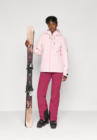 Világos rózsaszín sídzseki kapucnival, bordó sínadrággal és fekete síbakancsokkal. Fekete, mintás síléceket tart a kötésekkel együtt. Átlagos, funkcionális kialakítás.