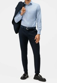 Světlomodrá košile na knoflíky s přední kapsou, tmavě modré kalhoty, texturovaný černý pásek a černé boty s masivní podešví. Sako přes paži.