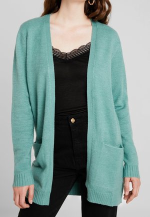 Femme portant un cardigan turquoise ouvert sur un caraco noir à bordure en dentelle et un jean noir taille haute.