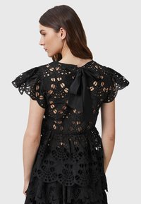 Black lace top featuring intricate cut-out patterns, ruffled short sleeves, and a bow at the back neckline; textured and layered design.