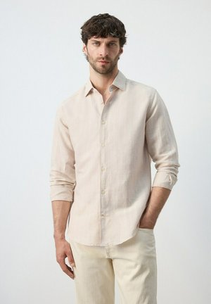 Hombre con cabello rizado oscuro, vestido con una camisa beige de manga larga abotonada y pantalones de color claro, de pie contra un fondo liso y claro.