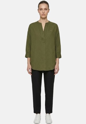 Donna in piedi, rivolta verso la fotocamera, che indossa una tunica verde oliva con scollo a spacco, pantaloni neri e sneakers bianche su uno sfondo semplice.