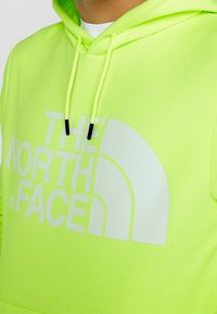 Ljus neon grön hoodie med en stor vit logo. Har snoddar med svarta spetsar. Mjuk tyg med en slät textur.