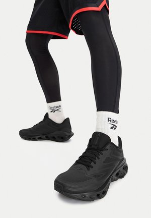 Osoba obutá v černých běžeckých botách, bílých ponožkách Reebok, černých legínách a černých šortkách s červeným lemováním, připravená k běhu.