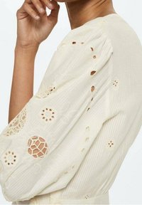 Cremfarbene Bluse aus strukturiertem Stoff, mit Puffärmeln und kunstvollen Spitzen-Ausschnitten, die florale Muster zeigen.