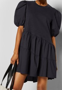 Navy blue puff-sleeve dress with a tiered hem, made from soft fabric, featuring a round neckline and gathered detailing. Held with a bag.