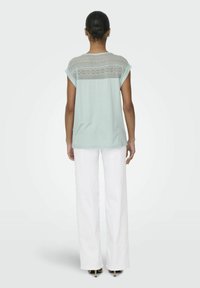 Jasno miętowa koszulka z koronkowym detalem na plecach, krótkie rękawy, zestawiona z białymi spodniami typu wide-leg i czarnymi szpilkami.