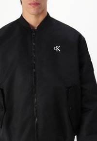 Giubbotto bomber nero realizzato in nylon liscio, dotato di zip frontale, tasche laterali e un logo ricamato bianco sul lato sinistro del petto.