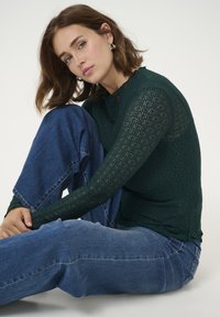 Top en dentelle vert foncé à manches longues, avec un motif texturé. Porté avec un jean en denim bleu clair, affichant une coupe décontractée.