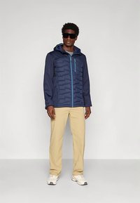 Navy jacket with a textured front and hood, featuring a light blue zipper. Paired with beige pants and white athletic shoes.
