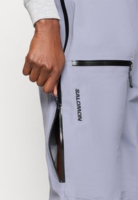 Svetlo vijoličast zip-up zunanji plašč z črnimi dodatki, ki ima stransko zadrgo in z logotipom "SALOMON" vezena spredaj v črni barvi.