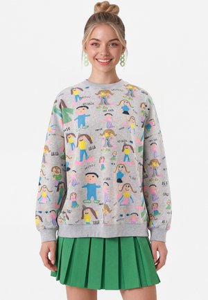 Ung kvinde smiler iført en grå sweatshirt med farverige, barnlige tegninger og tekst, kombineret med en grøn plisseret nederdel.