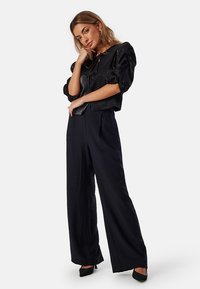 Top nero con maniche a sbuffo in tessuto testurizzato, abbinato a pantaloni neri a gamba larga. I tacchi completano l'outfit, evidenziando una silhouette elegante.