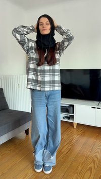Grå og hvid ternet skjorte, løse lyseblå jeans med frynser i kanten, sort tørklæde og mønstret sneakers, står på trægulv.