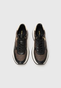 Een paar bruine en zwarte sneakers met zwarte veters, gouden oogjes, een beige hielaccent en een platform met witte zool op een lichte achtergrond.
