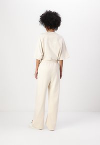 Lys beige bomullsdrakt med kortermet topp med preget tekst og bredbukser med elastisk midje. Enkelt design, myk tekstur.