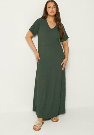 Frau steht und trägt ein dunkelgrünes, strukturiertes Maxikleid mit kurzen Ärmeln und geflochtene beige flache Sandalen vor einem einfarbigen hellen Hintergrund.