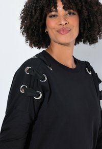 Černá mikina s kovovými očky na ramenou a tkanicemi z látky. Hladká textura, volný střih, kulatý výstřih.