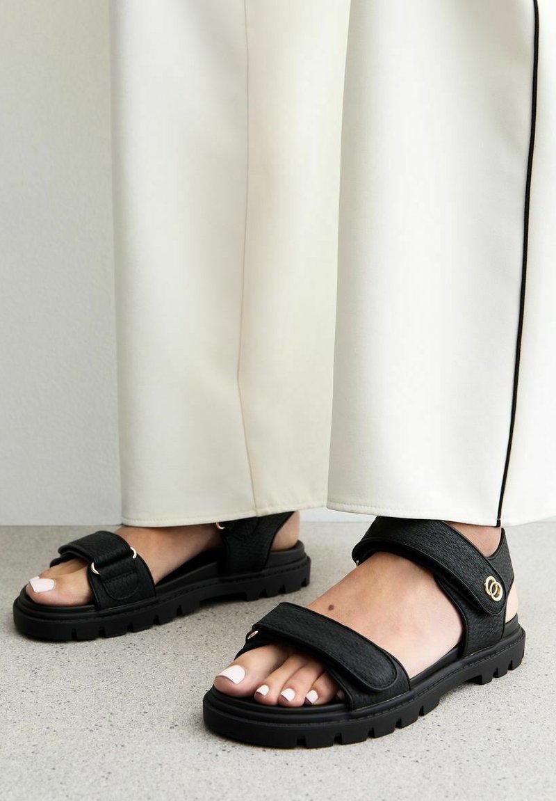 Svarta sandaler med texturerade remmar, justerbara kardborrestängningar och en tjock, robust sula. Vita lackade naglar synliga.