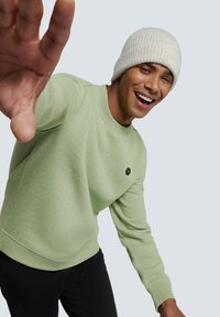 Lichtgroene sweatshirt met een ribbelnek, voorzien van een klein zwart hexagonaal logo, gecombineerd met een lichtgrijze gebreide beanie.