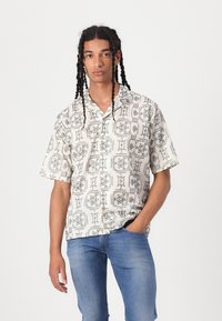 Νεαρός άνδρας με μακριά πλεξίματα, φορώντας λευκό πουκάμισο με σχέδιο και μπλε τζιν, στέκεται με το ένα χέρι στην τσέπη.