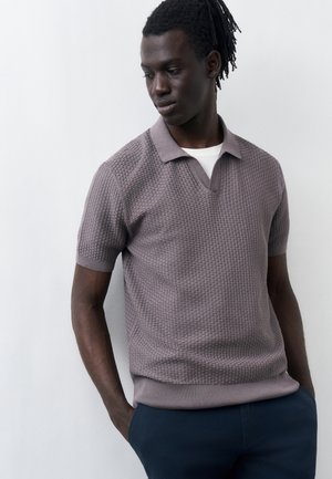 Fiatal, sötét bőrű férfi dreadlocksszal, szürke, texturált rövid ujjú galléros ingben és sötét nadrágban, kezét zsebre dugva, fehér háttér előtt.