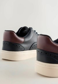 Ādas sporta apavi ar tumši ziliem un brūniem akcentiem, izceļas ar caurumu dizaina detaļām, apaļām auklām un baltu teksturētu zoli.
