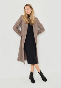 Long brown wool-blend coat with four buttons, worn over a fitted black ribbed dress and black ankle boots, against a neutral background.