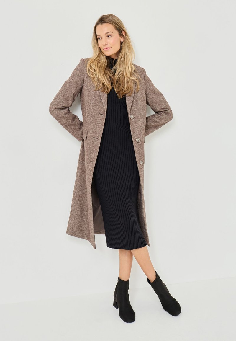 Long brown wool-blend coat with four buttons, worn over a fitted black ribbed dress and black ankle boots, against a neutral background.